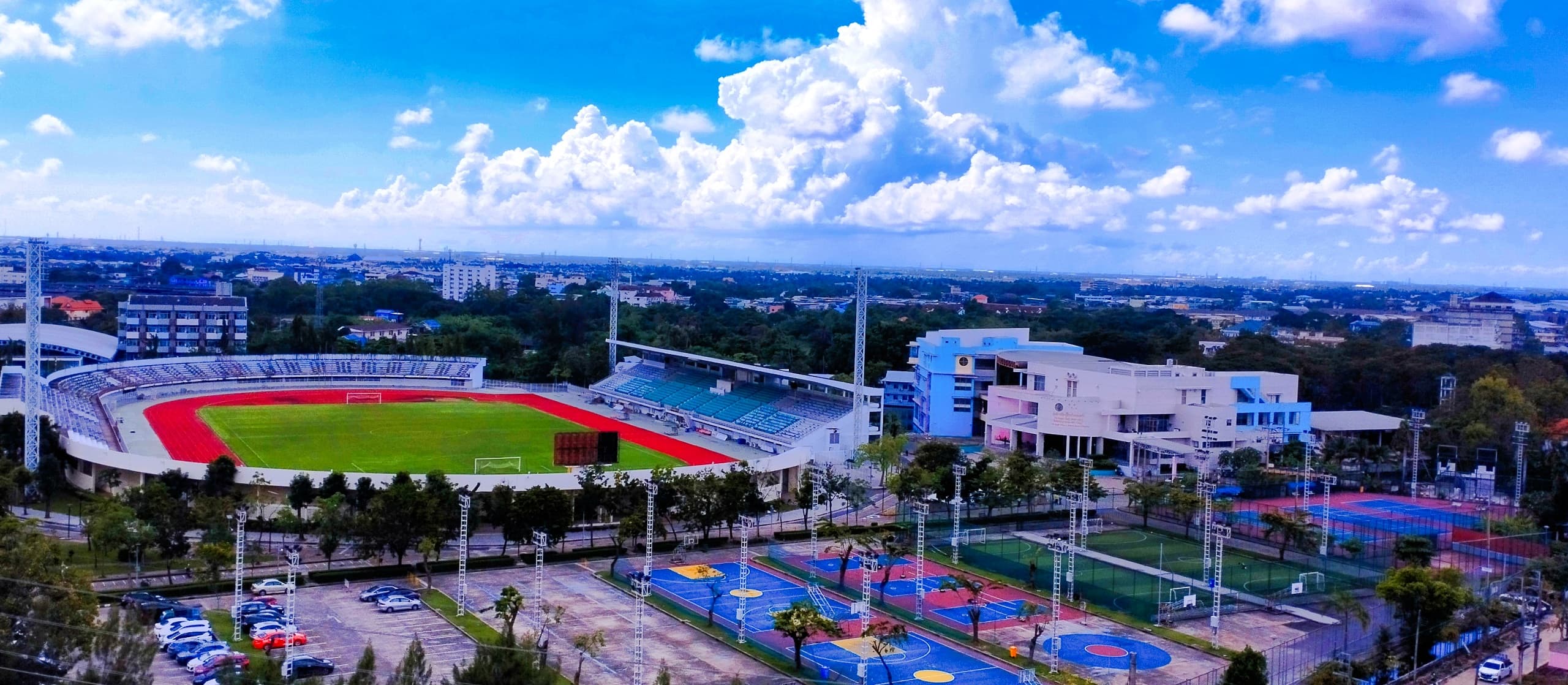 Aerial view of a campus sports complex with a red running track, outdoor courts, and a bright blue sky