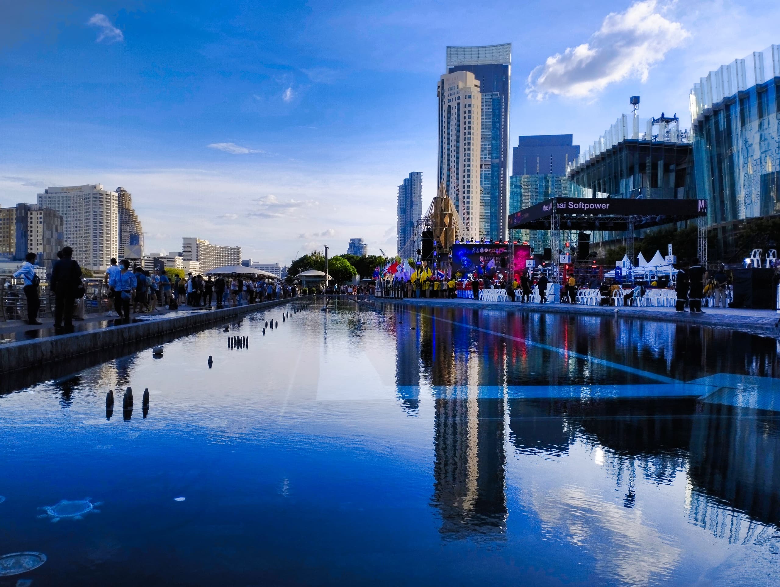 Reflecting pool mirroring Bangkok skyscrapers at dusk beside a waterfront promenade and event stage